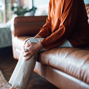 Elder person sitting on a brown leather couch, wearing a long-sleeved rust-colored top and light pants, holding one knee with both hands as if experiencing discomfort