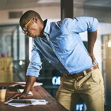 Man wearing a light blue button-up shirt and beige pants, leaning on a desk with one hand while holding the lower back with the other, suggesting back discomfort. Papers and a coffee cup are on the desk in an indoor office setting