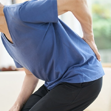 Person wearing a blue short-sleeved shirt and dark pants, leaning forward slightly while holding the lower back with one hand, suggesting back pain or discomfort. Indoor setting with soft lighting