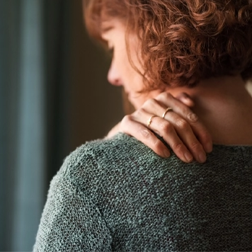 Woman wearing a textured green top, seen from behind, with one hand resting on the back of the neck, suggesting neck discomfort. The background is softly lit with a curtain visible